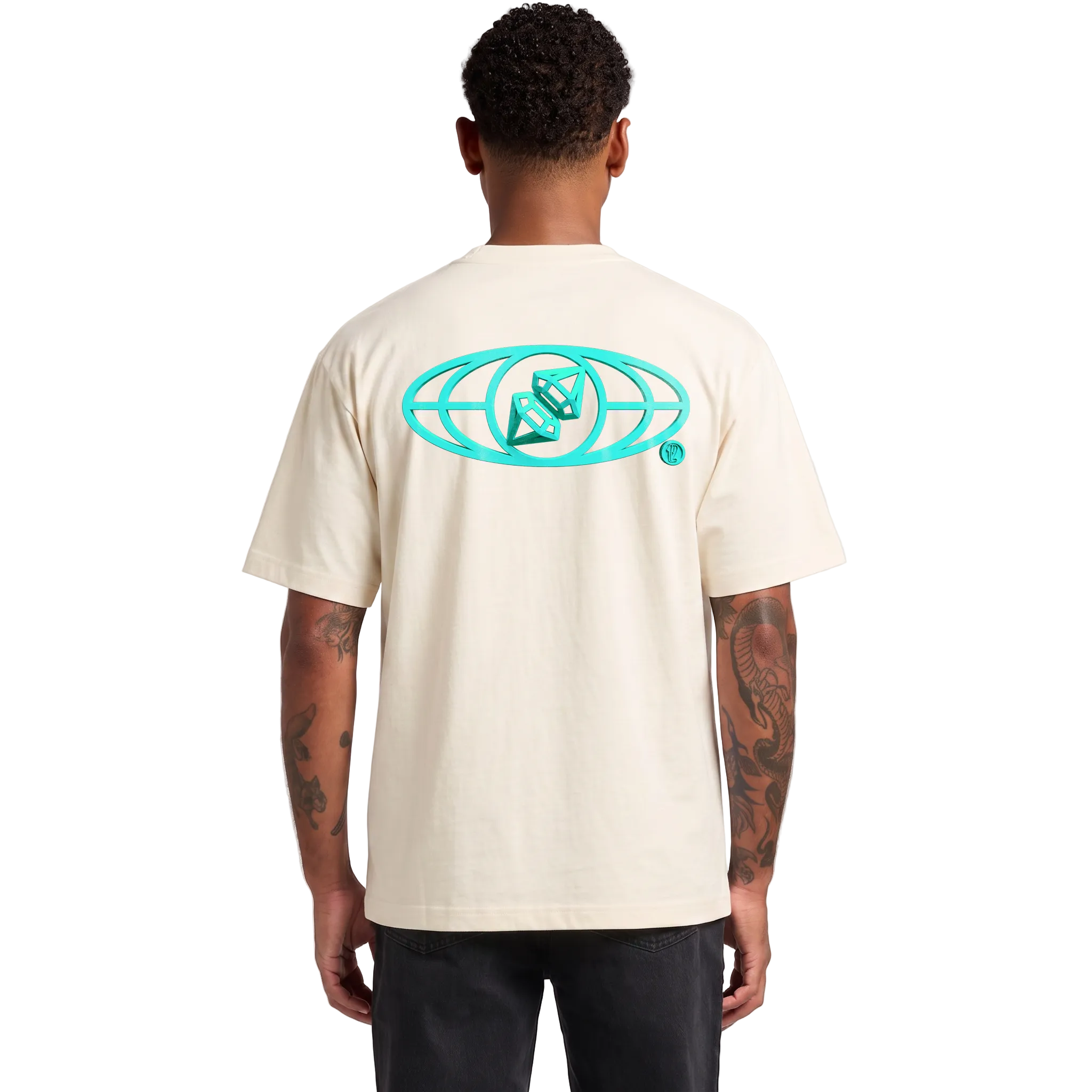 A man viewed from the back wearing a cream-colored t-shirt with a large teal graphic on the back, paired with black pants, against a light gray studio background.