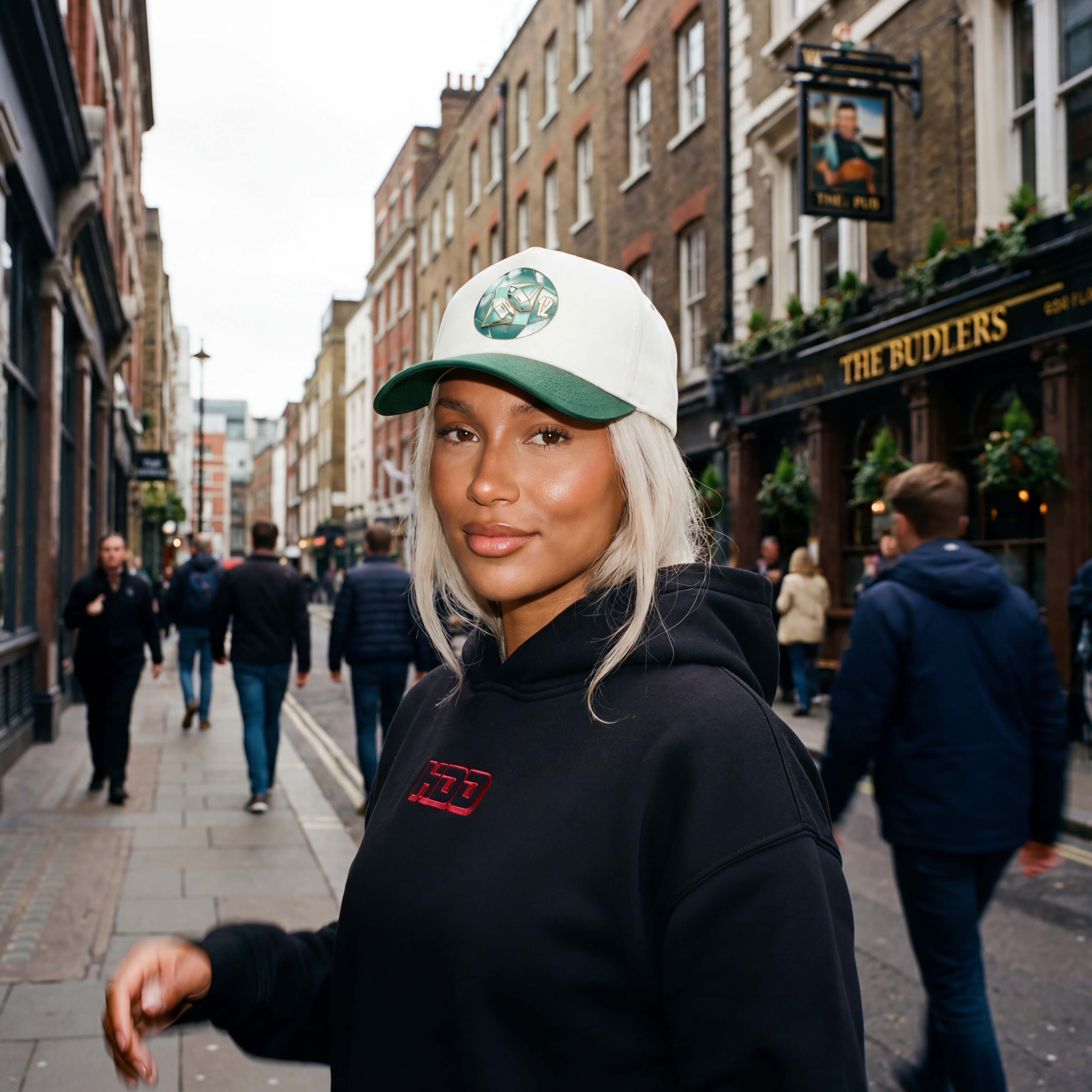 Woman wearing a black hoodie with a logo and a white cap with a green logo on a city street.
